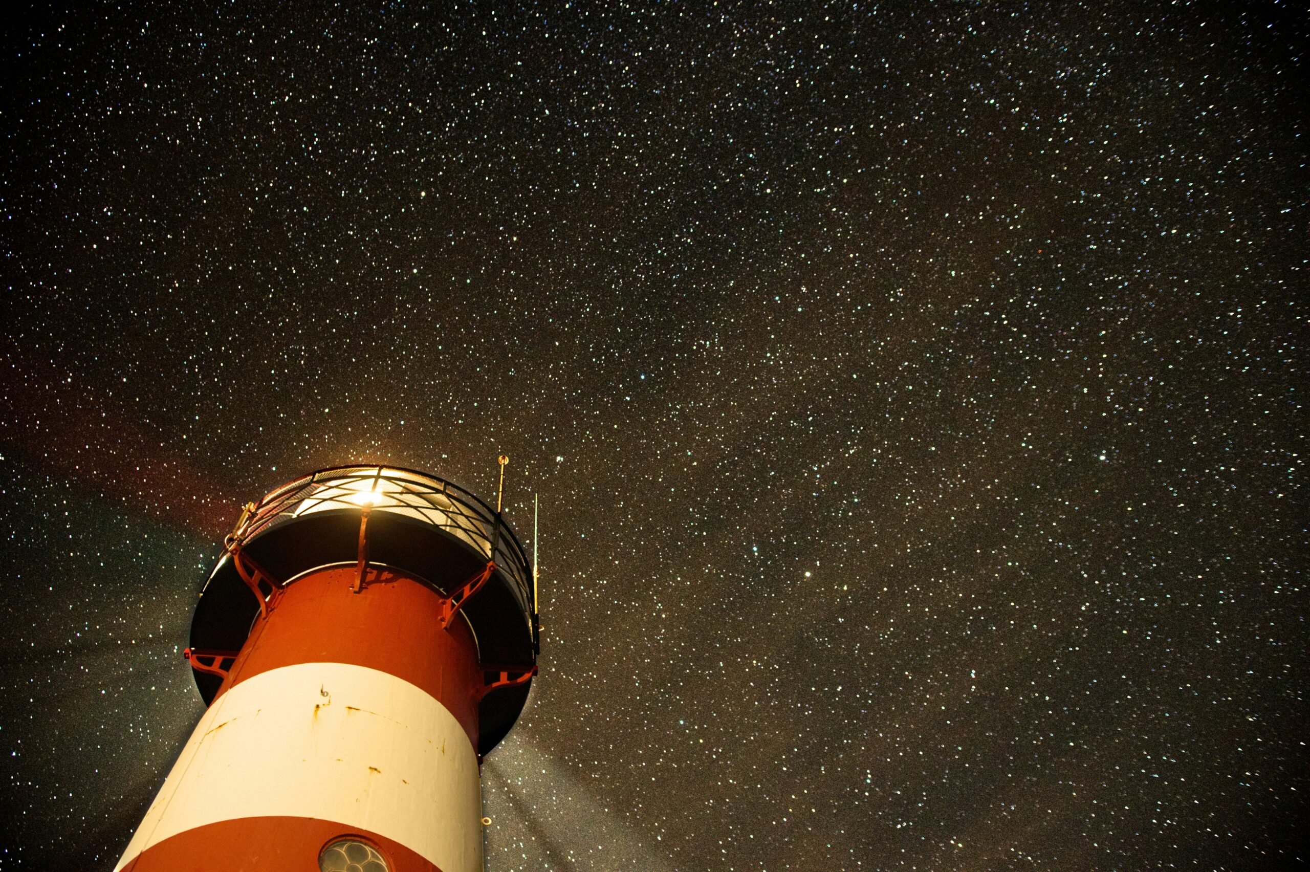 A red and white lighthouse against a background of the night sky, filled with stars.