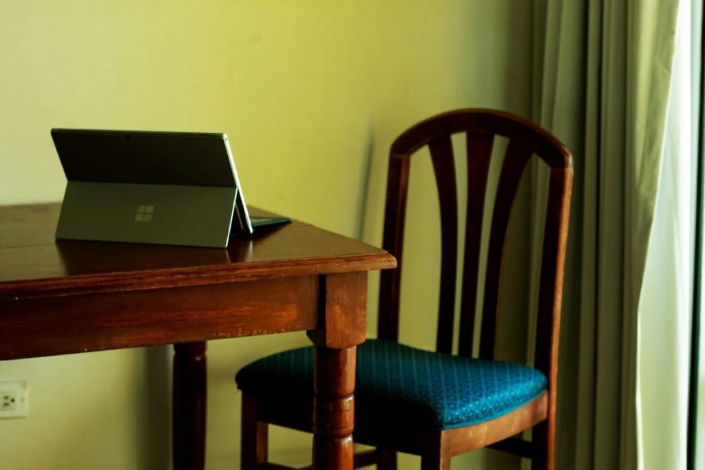 An open tablet computer sits on a wooden table before an empty wooden chair with a blue cushion.