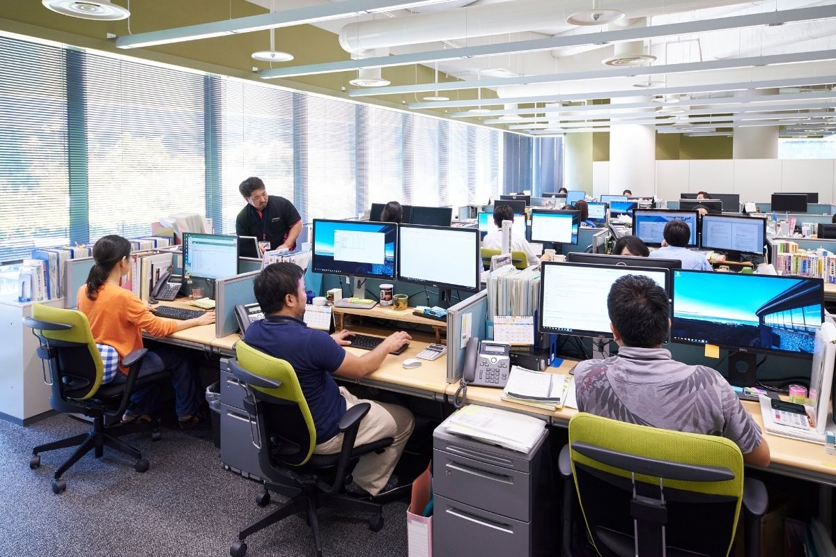 At an office, people are at multiple workstations in rows of three people all looking at computer screens in front of them.